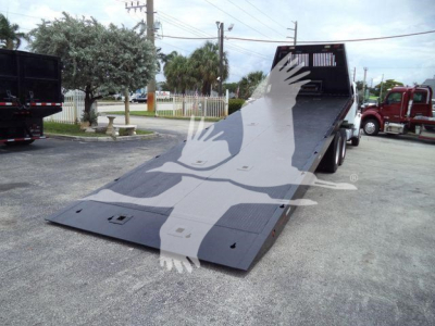 Rolloff truck with a lowered metal ramp platform in a truck yard. Flatbed/deck shows non-slip surface and two rear wheel areas; Freightliner M2 106 likely. No visible text readable on decals.