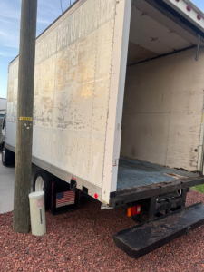 Box truck with open rear doors showing cargo area; faded lettering on trailer walls, rural ground with gravel and utility pole nearby.