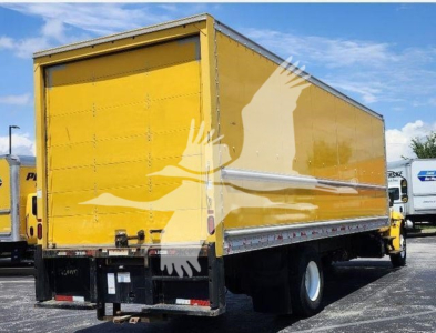 Box truck rear/side view showing yellow cargo box with roll-up door and black bumper; International MV 2021 used truck in background.