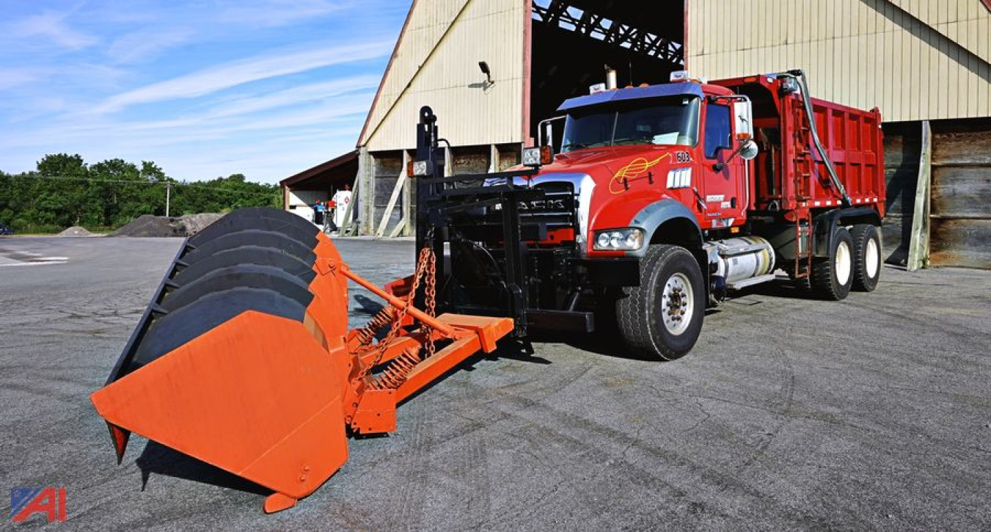 Truck with orange snow plow and spreader attachment in front of a red Mack GU713 Granite dump truck under a hangar.