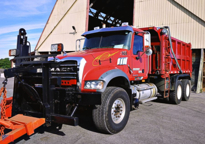 Mack GU713 Granite dump truck with plow and spreader attachment, red cab and dump body, side angle view.