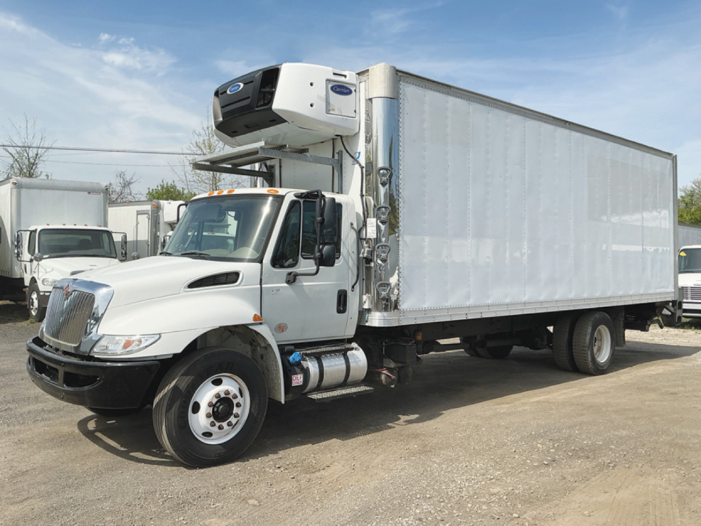International 4300 refrigerated box truck with a mounted refrigeration unit on the cab, white cab and silver cargo box, parked outdoors.