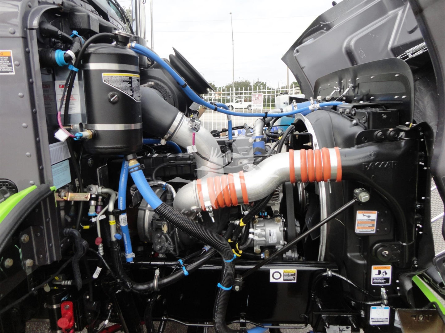 Engine bay of a 2022 Peterbilt 337 with visible blue and orange hoses, intake piping, and various hydraulic lines.