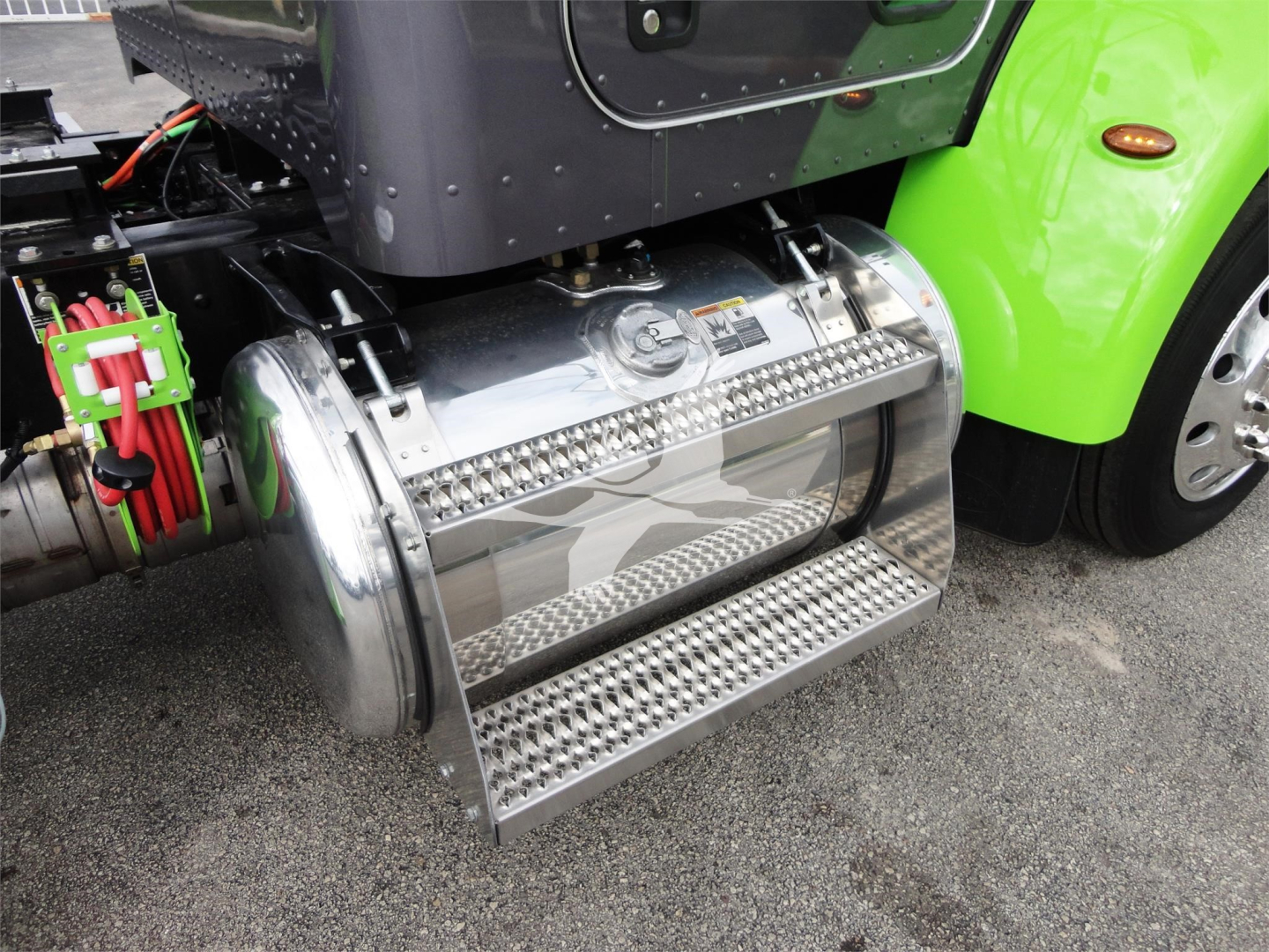 Truck fuel tank with polished chrome cylindrical tank and metal diamond-plate steps mounted under a bright green Peterbilt cab.