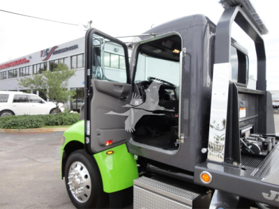 Peterbilt 337 rollback truck with open cab door showing interior, black chassis, and hydraulic bed; bright green fender and trailer in background.