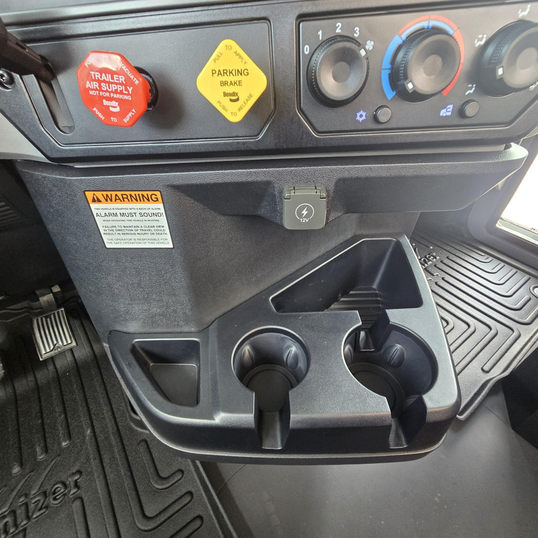 Cab interior dash area of a 2027 Mack PI64T showing parking brake yellow knob, red trailer air supply knob, cup holders, and control panel with climate dials.
