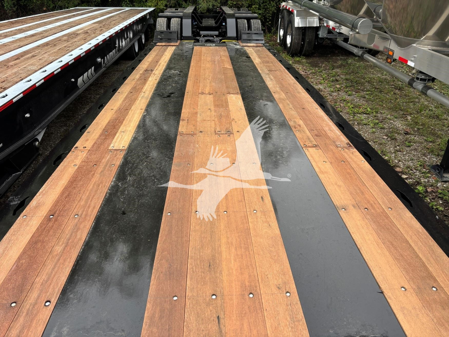 Flatbed trailer deck with wooden planks and black steel strips, viewed from end near the gooseneck, surface shows natural wood and sealed areas.