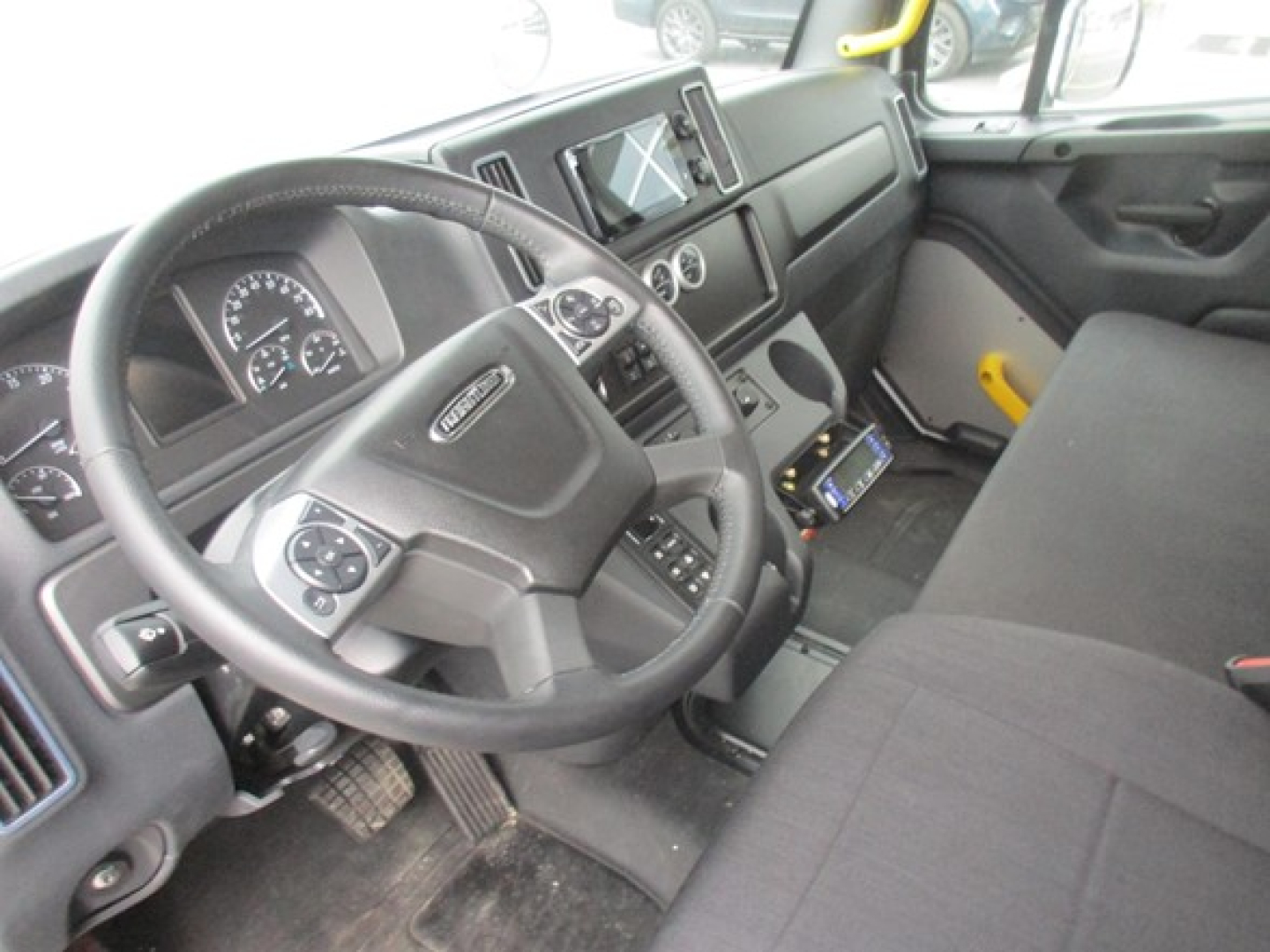 Truck cockpit showing steering wheel, instrument cluster, center console with digital display, and driver-side door; Freightliner branding on steering wheel interior.