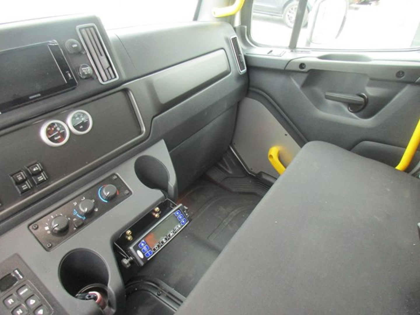 Truck cabin interior showing dashboard with steering wheel on left, radio touchscreen, climate controls, two circular gauges, and a center console with a device mounted; passenger seat visible.