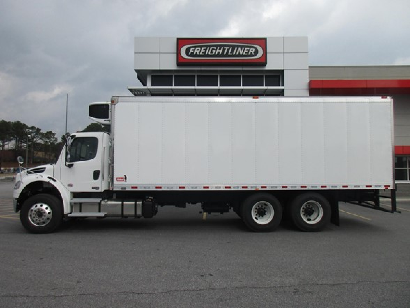 Freightliner truck in front of a dealership building with a large refrigerated box body, side profile view showing three rear axles and white cargo trailer.
