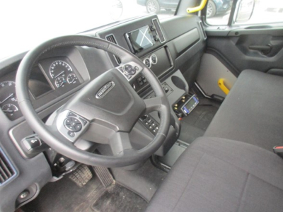 Truck cockpit showing steering wheel, instrument cluster, center console with digital display, and driver-side door; Freightliner branding on steering wheel interior.