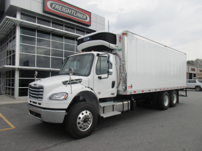 Freightliner M2 106 Plus refrigerated truck with white cab and box, parked in front of dealer building with Freightliner sign.