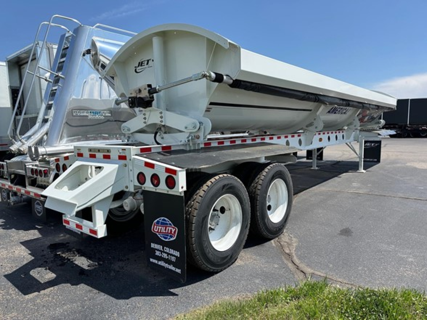 Jet side-dump trailer with dual-axle wheels and white body, rear loading chute, reflective strips, and hydraulic hoist mechanism visible.
