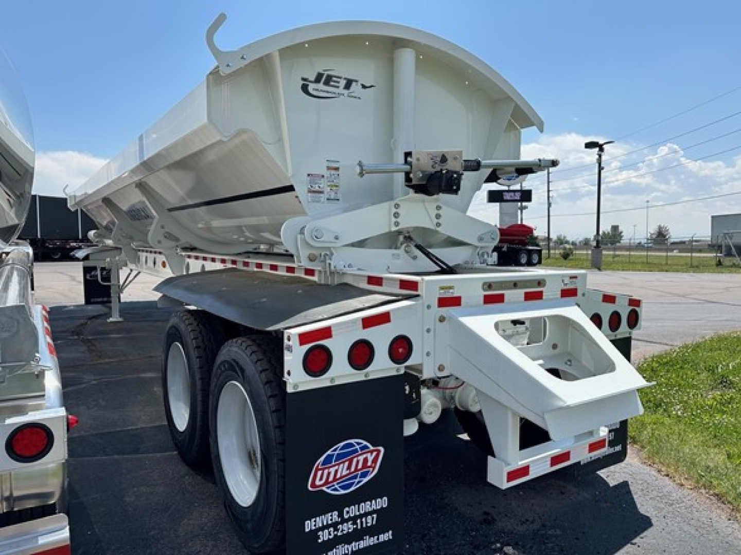 Jet Jet side dump trailer showing rear view with white body, dual rear axles, and utility sticker. Visible hydraulic mechanisms and mounting bracket on the front of the dump body.