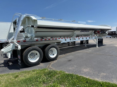 Dump trailer in side-dump configuration showing tandem axle wheels and white painted steel body with American made branding and safety rails.