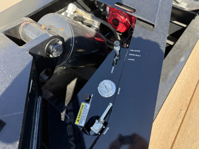 Spreader bar attachment on a trailer with hydraulic ram and control levers visible, including caution label and red motor housing.