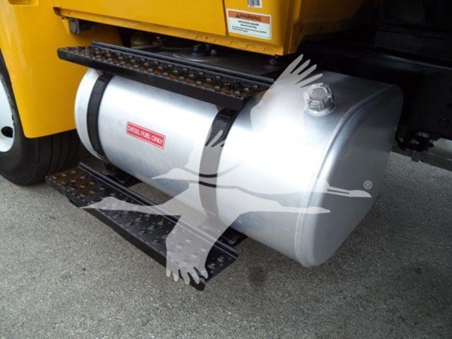 Fuel tank mounted under a yellow International Durastar 4300 truck showing a silver cylindrical fuel tank with a red label, overhead steps, and part of a black frame.