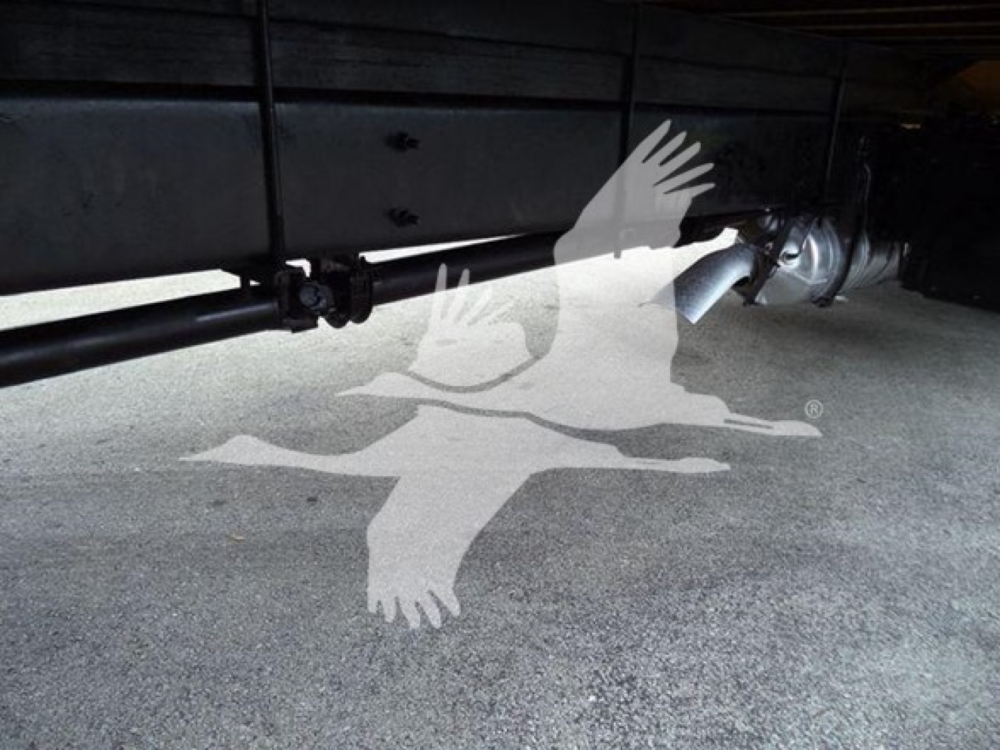 Vehicle undercarriage showing trailer hitch and exhaust pipe with shadowed logo overlay.