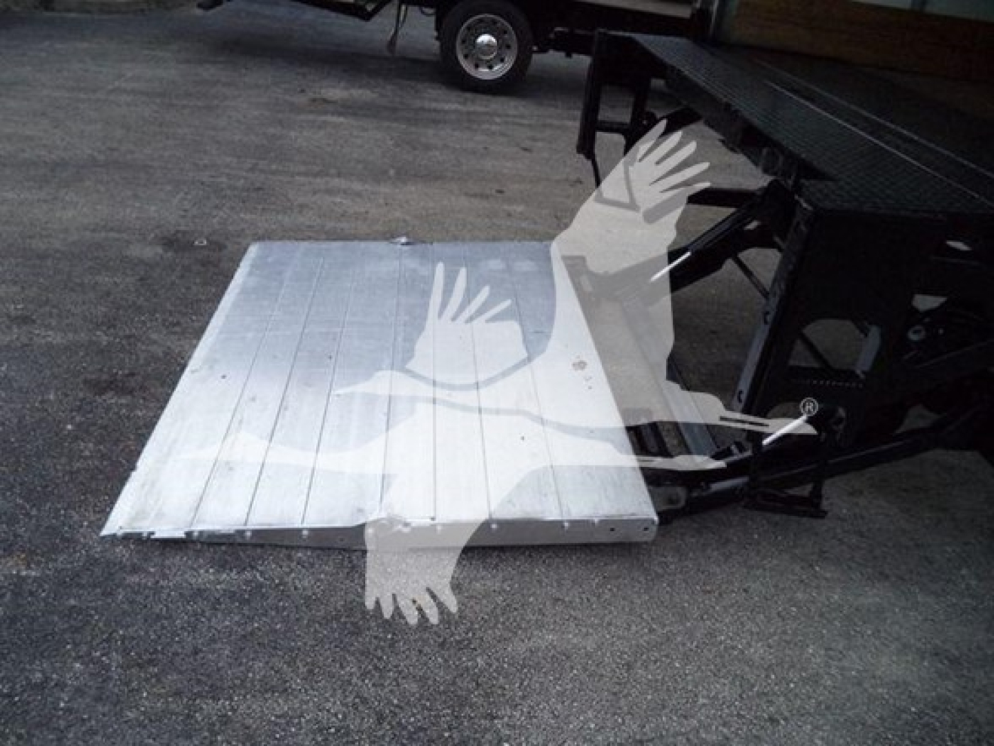 Platform with aluminum ramp or plate on ground near a truck chassis; visible text not legible due to watermark; overall shows a metal plate and part of a vehicle frame.