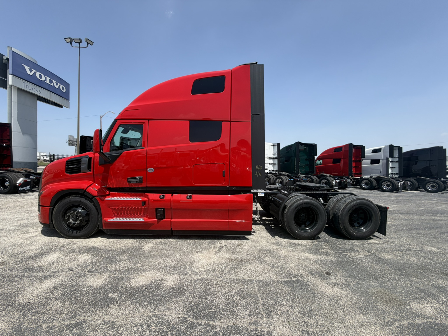 Truck side profile showing a red 2027 Mack PR64T76H sleeper cab tractor with a long hood, two side windows, and a black chassis with triple rear axles.