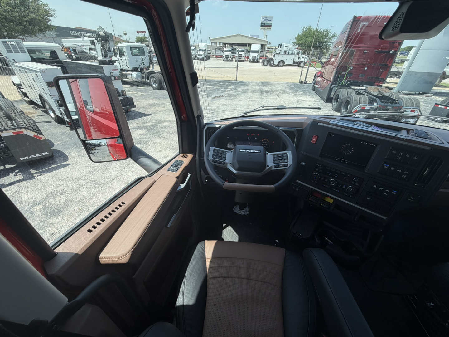 Truck cab interior with steering wheel, dashboard, and door panel visible; window shows yard with trucks and trailers.