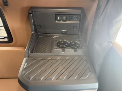 Interior truck cab area showing center console with storage and power outlets in a Mack PR64T76H. Visible features include cup/phone holders, a rectangular power outlet panel, and textured fold-out surface.