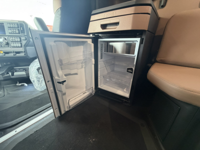 Cab interior showing a small fridge/freezer in the dash area of a Mack PR64T76H truck, with open mini-refrigerator door exposing empty shelves and interior.