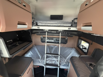 Truck sleeper cab interior showing bunk bed with ladder, storage compartments, and plastic-covered mattress; beige and black trim with side windows.