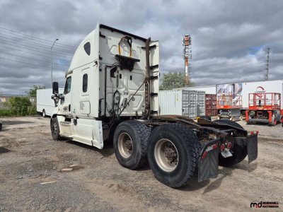Freightliner Cascadia tractor rear/side view showing chassis and cab with fifth wheel mount removed; notations on cab and mudflaps, multiple tires, and parking lot background.