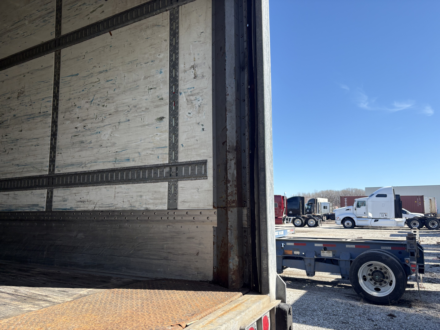 Trailer interior showing roll door opening with metal side rails and wooden wall, open bay with blue sky outside.