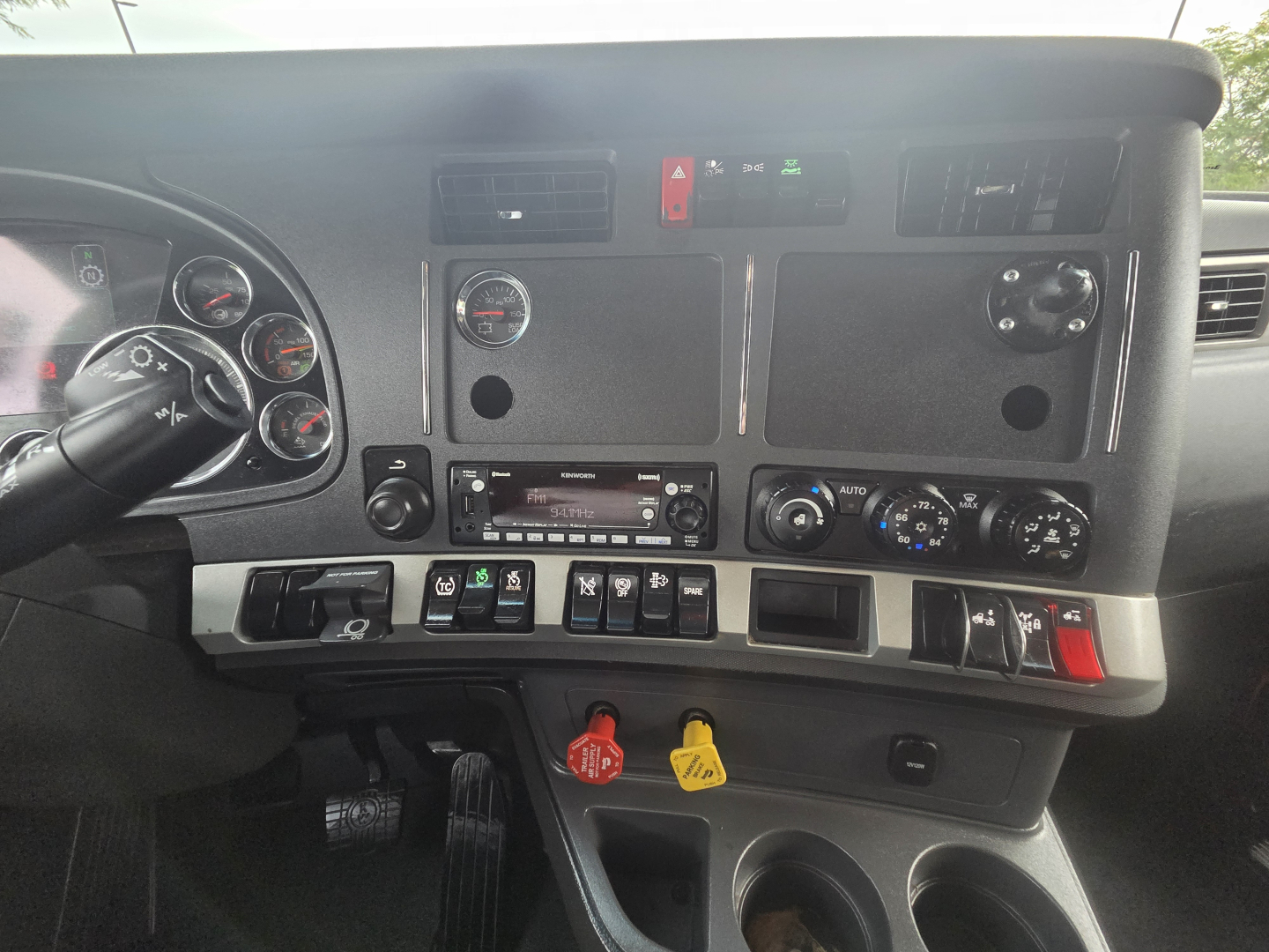 Kenworth T680 dashboard and center console. Dashboard gauges, switches, HVAC controls, audio head unit, and red/green indicator lights visible. Includes steering wheel and pedal area partially visible.