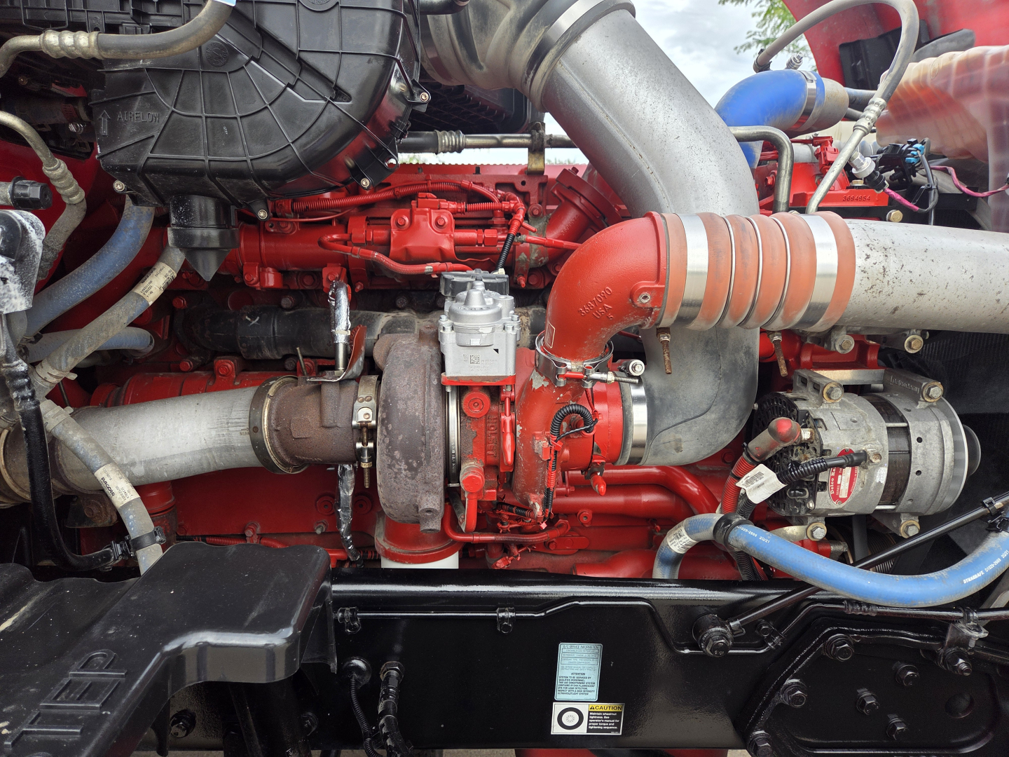 Kenworth T680 engine bay showing red diesel engine with air intake piping and turbocharger, blue hoses, and various metal clamps.