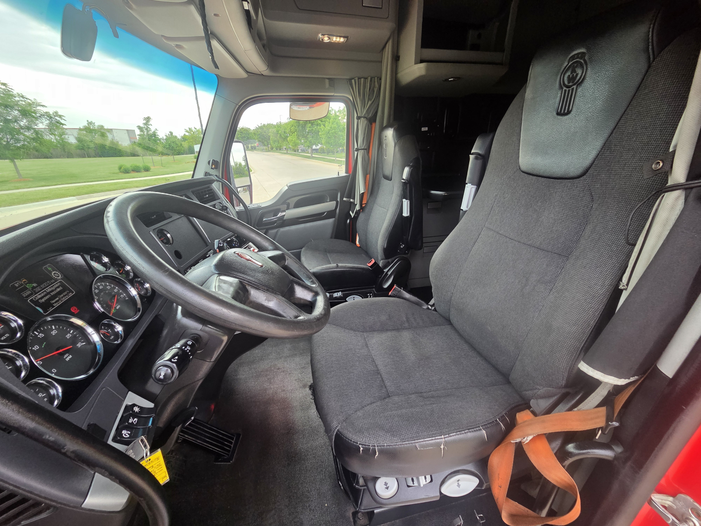Kenworth T680 cab interior showing steering wheel, dashboard gauges, and driver seat with seat belt in view.