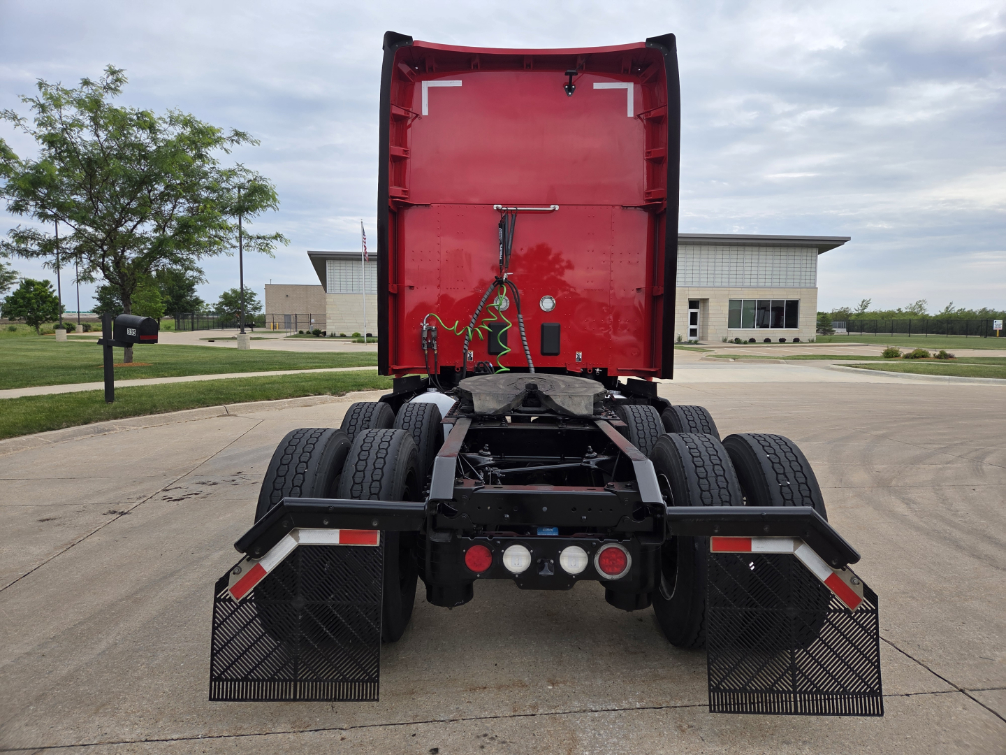 Kenworth T680 rear view showing fifth wheel and dual rear axles with mud flaps and wiring, red cab exterior, clean truck yard.