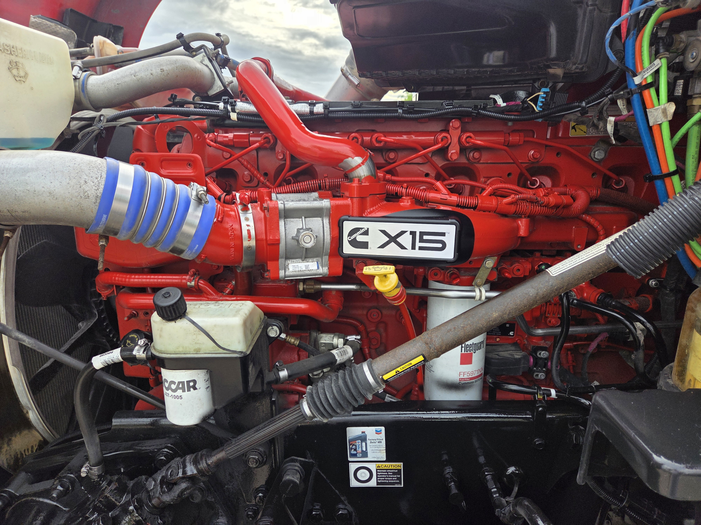 Kenworth T680 engine bay with red engine block and blue hose, label X15 on intake, various hydraulic lines and reservoirs visible.