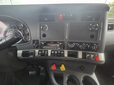 Kenworth T680 dashboard and center console. Dashboard gauges, switches, HVAC controls, audio head unit, and red/green indicator lights visible. Includes steering wheel and pedal area partially visible.