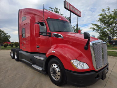 Kenworth T680 sleeper cab truck in red, side view showing large chrome grille, dual rear axles, black bumper, and Kenworth dealership sign in background.