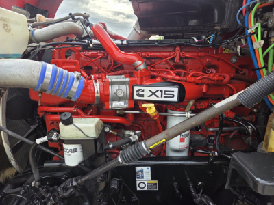 Kenworth T680 engine bay with red engine block and blue hose, label X15 on intake, various hydraulic lines and reservoirs visible.