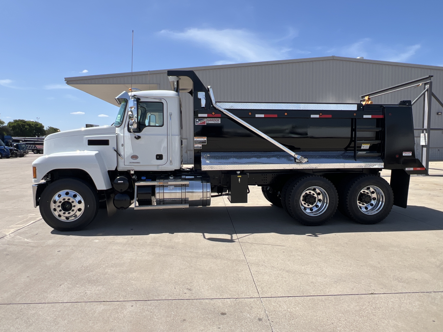 Dump truck showing 2027 Mack PI64 with white cab and black dump body, side view in parking lot.