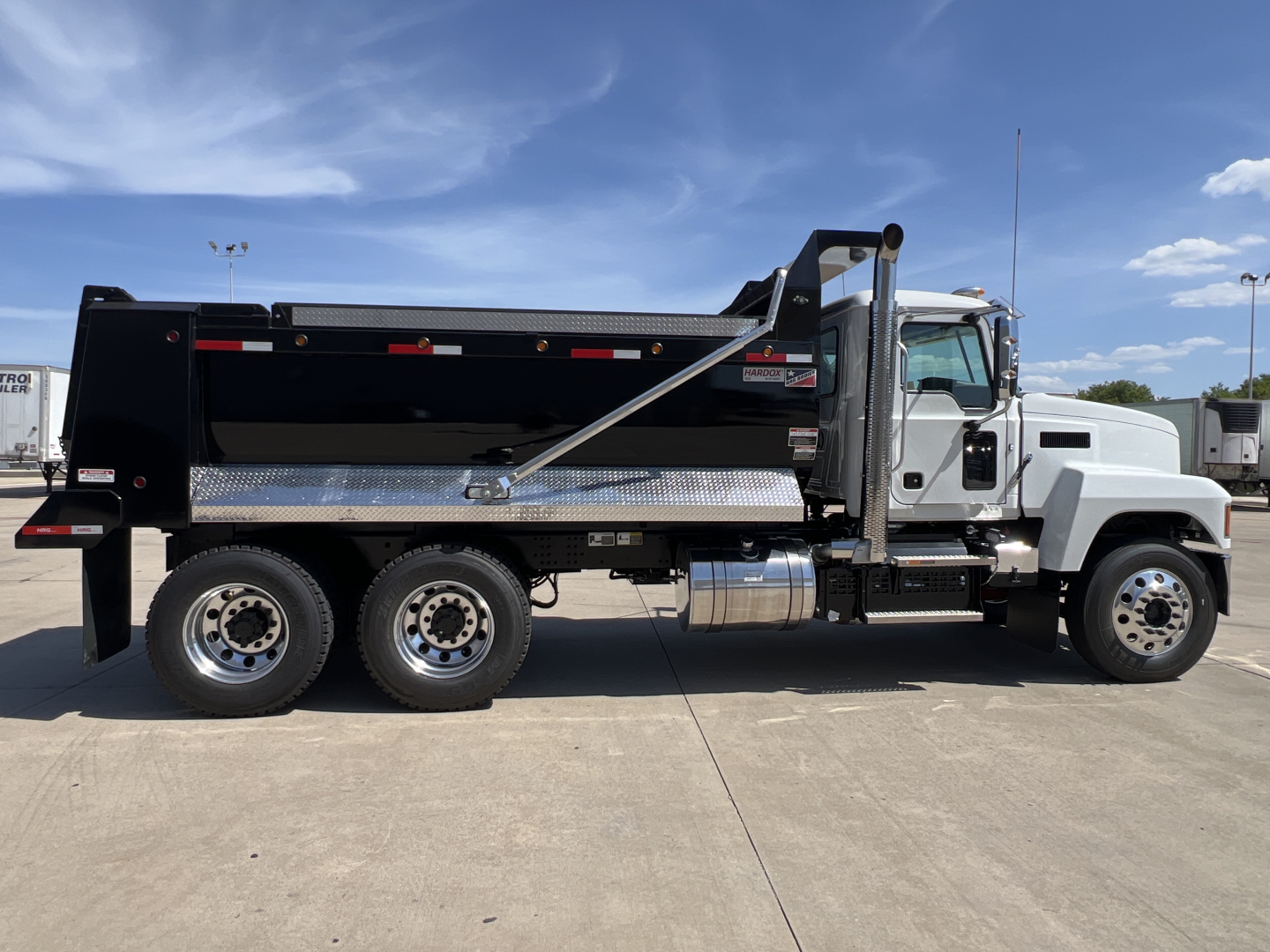Side view of a 2027 Mack PI64 dump truck showing black dump body, chrome fuel tanks, and dual rear axles with six wheels.