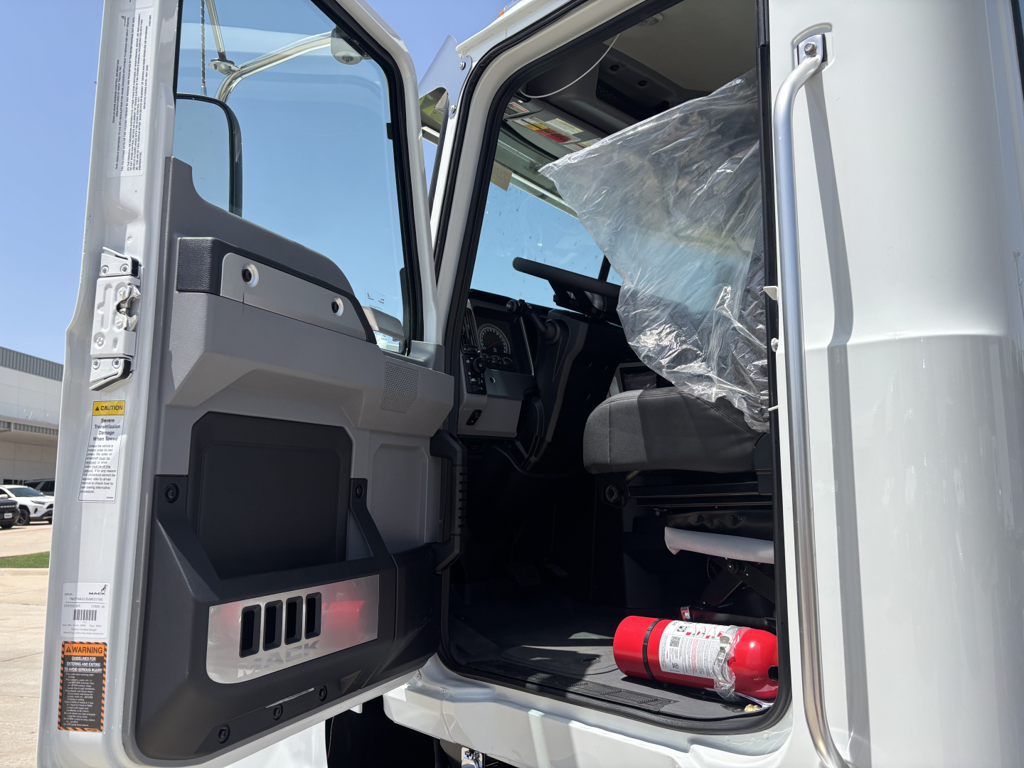 Cab interior of a 2027 Mack PI64 dump truck showing open driver door, dashboard, steering wheel, and plastic-wrapped seat; fire extinguisher in cab.