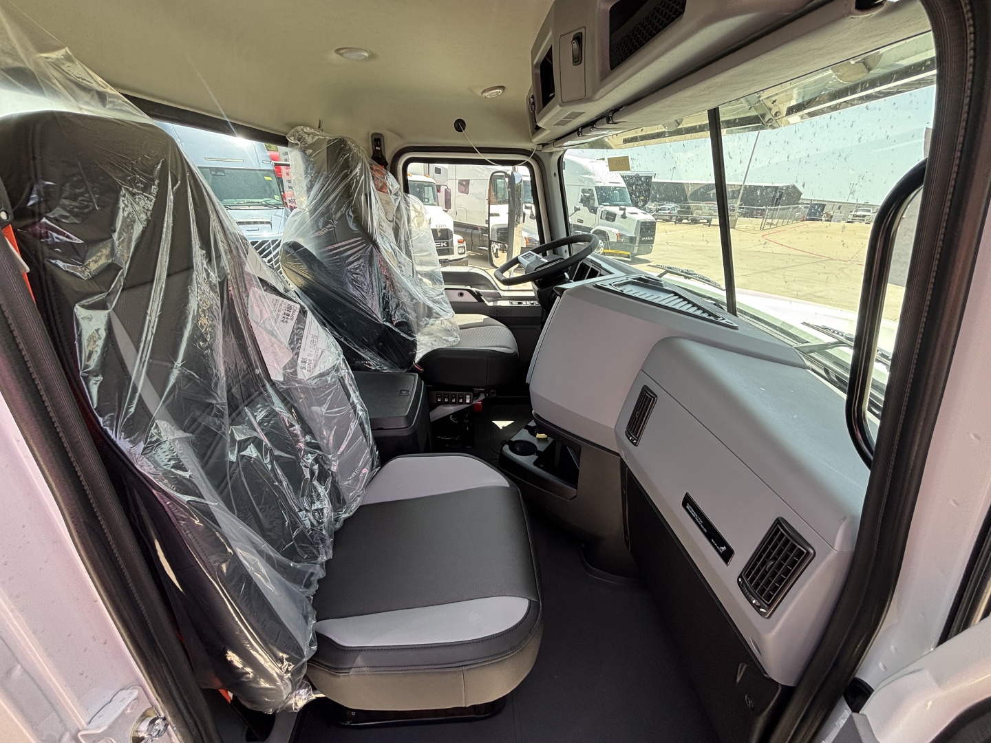 Interior cab of a new 2027 Mack PI64 dump truck showing front bench seat with plastic covering, steering wheel, dashboard, and large windshield.