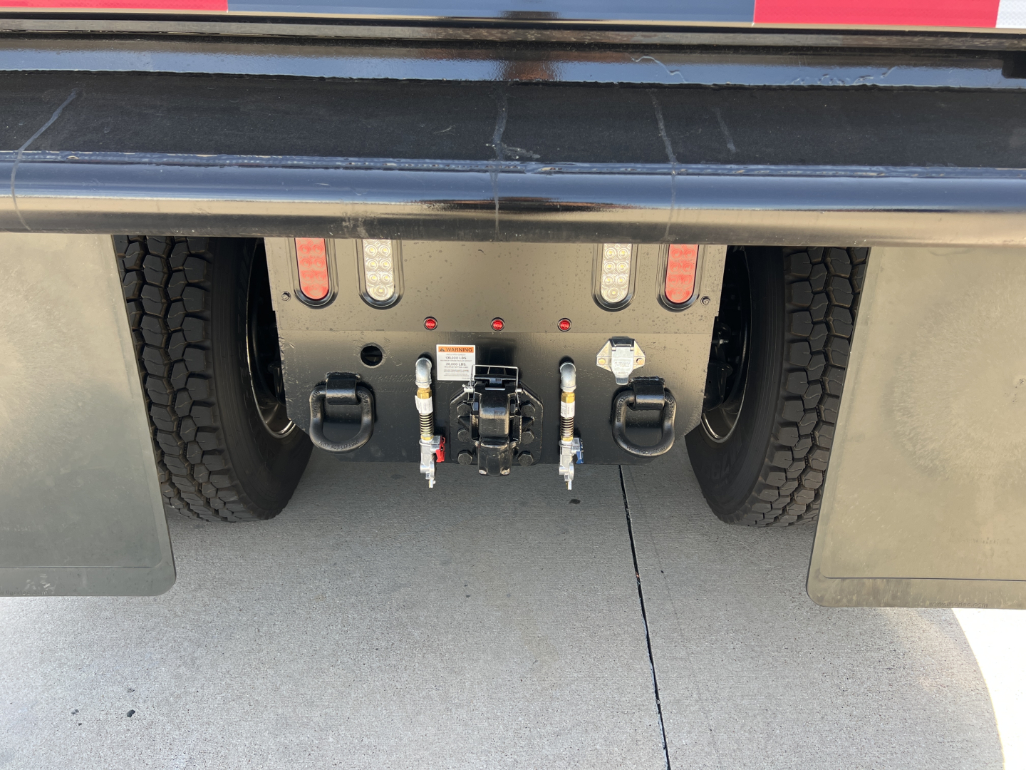 Rear view of a Mack PI64 dump truck chassis showing the towing and hitch connection with hoses and electrical connectors, rear tires on each side and mudflaps.