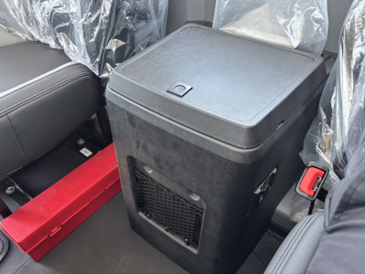Dump truck cab interior showing a black portable cooler or storage unit in middle with plastic seat coverings, red tool/box, and seatbelt buckle visible.