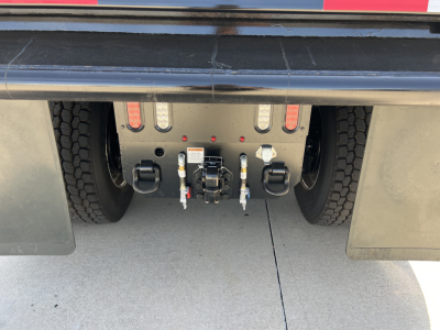 Rear view of a Mack PI64 dump truck chassis showing the towing and hitch connection with hoses and electrical connectors, rear tires on each side and mudflaps.