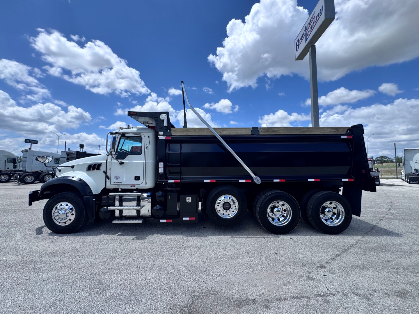 Dump truck side view showing Mack GR84F dump truck with black dump bed and white cab, three rear axles, and side rails.