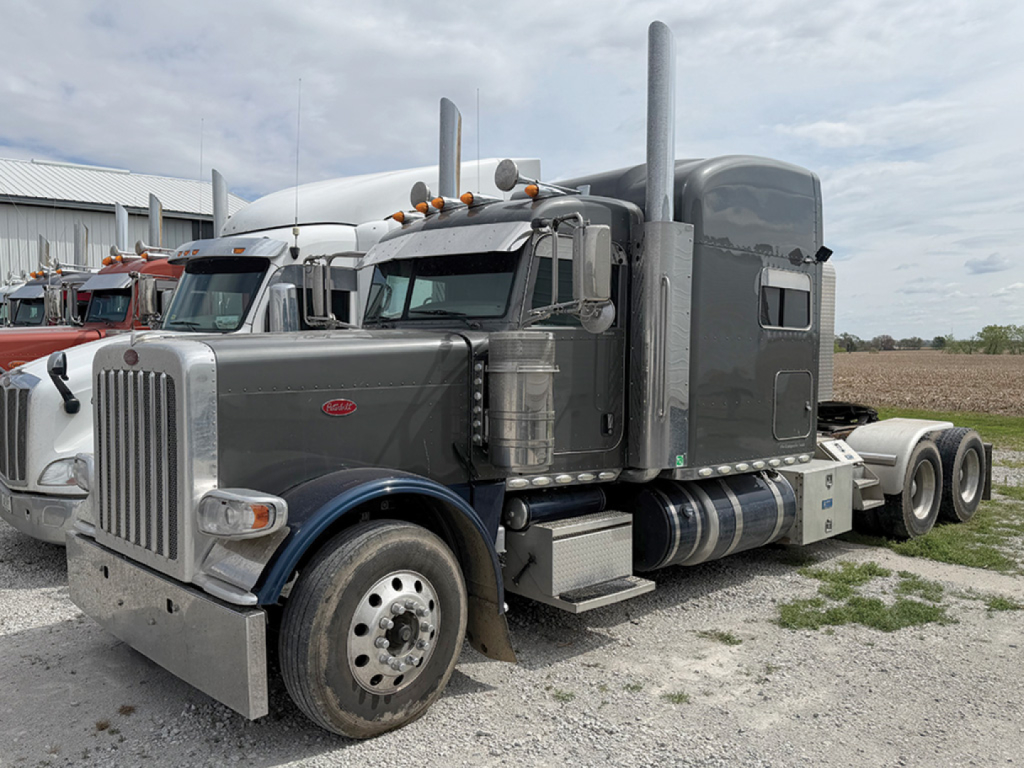 Peterbilt 389 UltraCab conventional Sleeper tractor in gray with tall sleeper and dual high exhaust stacks, front chrome grille, large round headlights, and dual rear axles on a gravel lot.