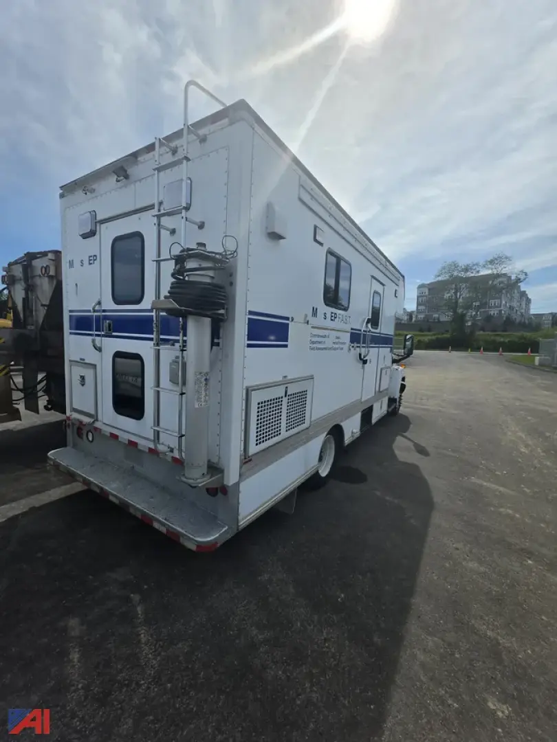 Mobile lab truck rear/side view showing exterior equipment and access doors; multiple windows; ladder; utility equipment mounted on rear; blue stripe branding.