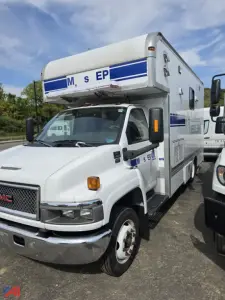 Truck cab in front shows GMC emblem and attached mobile lab housing with blue stripe graphics. Text on side reads M S EP or similar. Large side compartment doors, windowed cab, overhead sleeper unit.