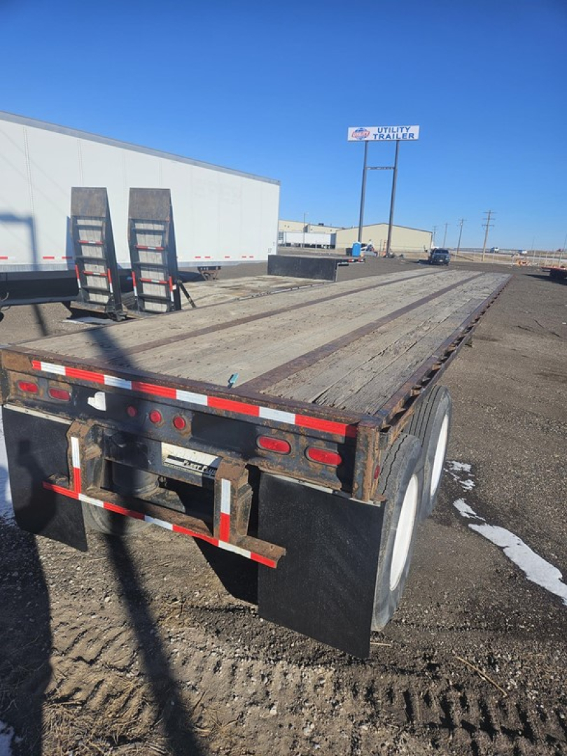 Flatbed trailer rear view showing wooden deck, dual tires, mud flaps, and rear lights. Partial stairs/ladder leaned on left side of trailer. Clear sky and storage yard in background.